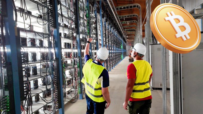 Two workers in high-visibility vests and hard hats walk through a Bitcoin mining facility with rows of computer hardware, while a large Bitcoin coin graphic appears in the upper right corner.