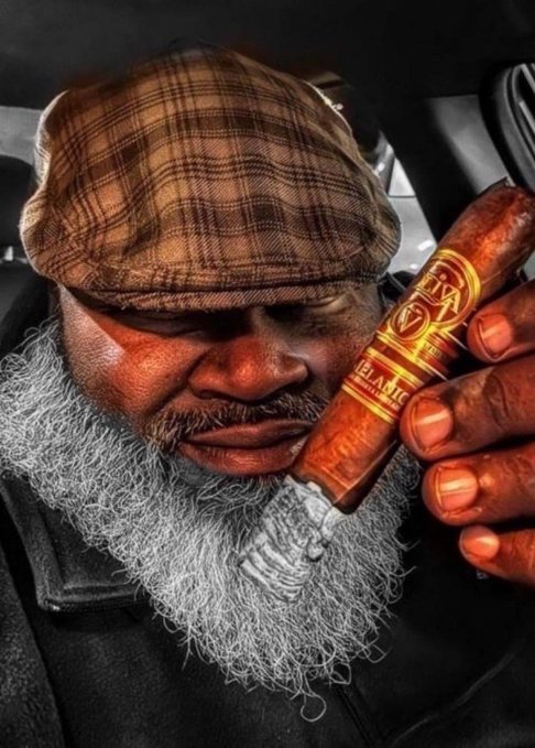 A close-up of an older man with a gray beard wearing a brown plaid flat cap, holding a large brown cigar near his face.