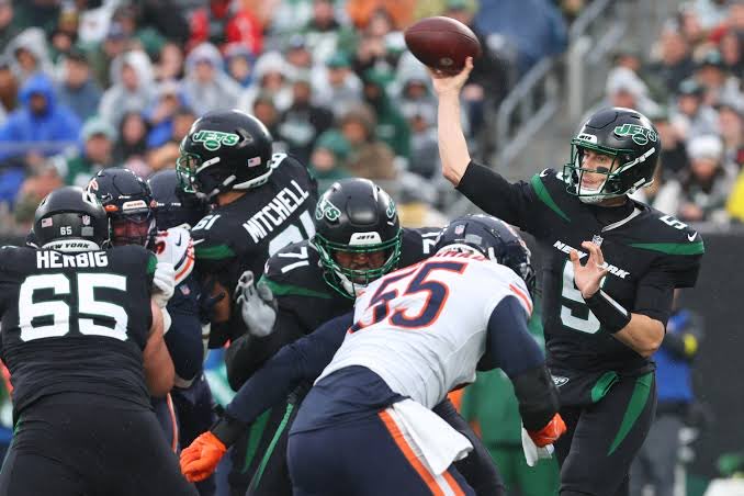 An NFL game action shot showing New York Jets quarterback throwing the football past Bears defenders, with player Hergig #65 and Mitchell visible.