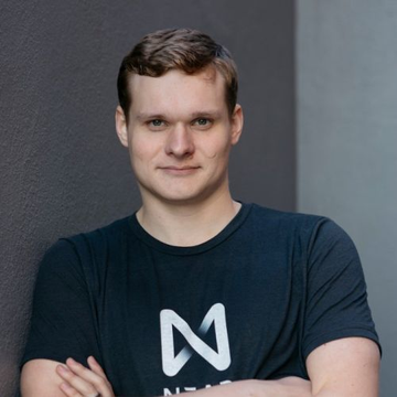 A portrait of NEAR co-founder Illia Polosukhin, wearing a black t-shirt featuring the NEAR Protocol "N" logo, accompanying the news that he advocates for verifiable AI infrastructure and open-source evaluation rather than slowing AI research.