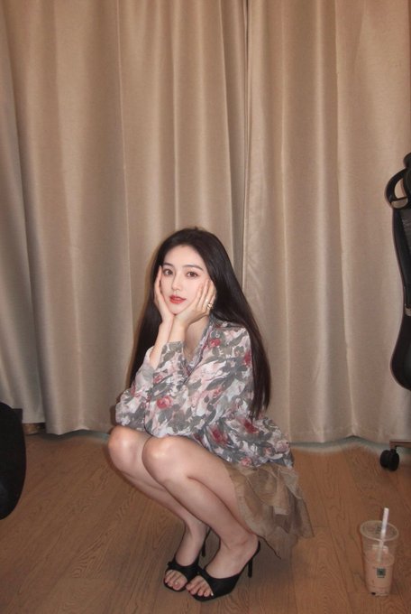 The image features a woman sitting cross-legged with her hands on her face, posing for a photo. She is wearing a floral dress that has a skirt, which adds an elegant touch to her appearance. The background of the picture includes a curtain and a chair, suggesting that she might be in a room or a dressing area. There are also two cups visible in the image - one on the floor near the woman's feet and another further away from her.