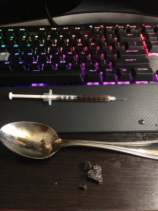 A darkened desk scene featuring an RGB backlit keyboard, a syringe, and a spoon with dark residue next to a small pile of substance.