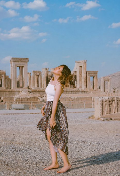 A woman in a white tank top and patterned skirt stands barefoot in front of ancient stone ruins under a bright blue sky, her head tilted back and hair flowing.