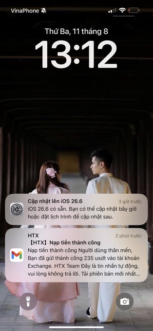 An iPhone lock screen screenshot from a VinaPhone carrier displaying the time '13:12' on Thursday, August 11, with two notifications about iOS 26.6 update and HTX Exchange topping up 235 USDT, over a wallpaper of a couple in traditional clothing.