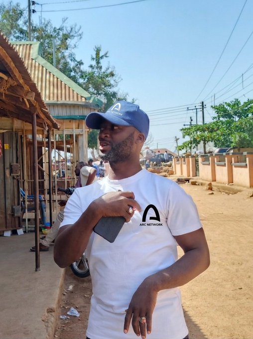 A man wearing a blue cap and a white t-shirt with "ARC NETWORK" branding stands outdoors in a sunlit street setting, posing to introduce himself as a member of the Arc (ARC) community. His relaxed posture and the branded apparel visually reinforce his role as an active community member ready to build, test, and create content on Arc.