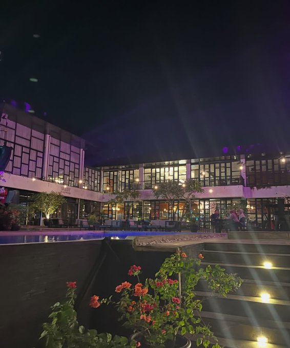 Nighttime photo of a stylish outdoor venue with a pool, illuminated stairs, and a multi-story building with lit windows, viewed past pink flowers in the foreground.