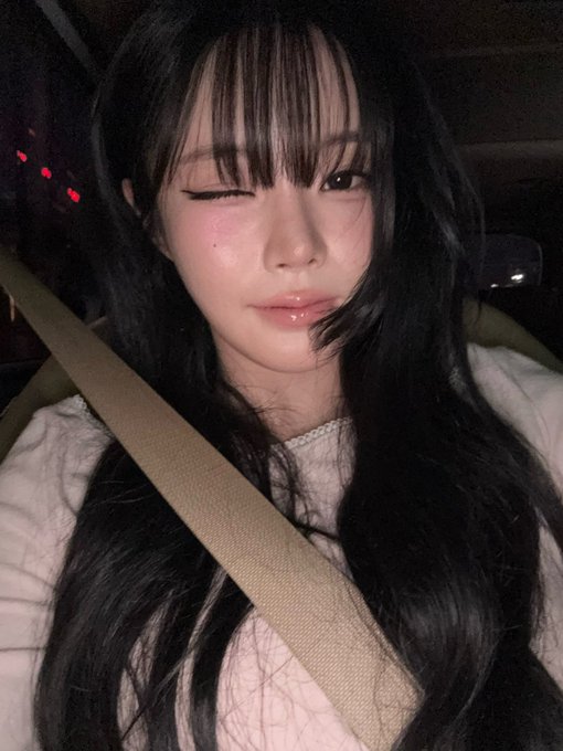 A young woman with long dark hair and bangs sits in a car wearing a seatbelt, winking and making a soft pout at the camera. The image accompanies a post about KEENA of FIFTYFIFTY, suggesting the photo features the K-pop idol sharing a candid update.