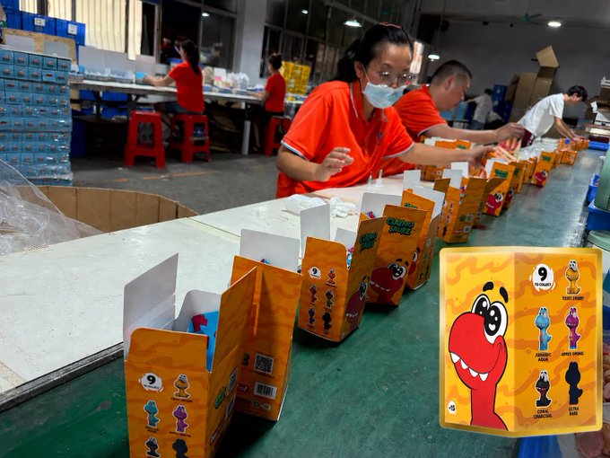 Workers assemble orange "Cardboard Games" blind boxes featuring a red dinosaur character and "9 designs" markings, preparing them for shipment to DYLI HQ.