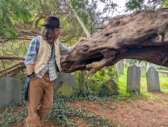 A man dressed in period clothing with a brown hat stands in a cemetery, touching the snout of a large, weathered tree root or branch sculpture that resembles an animal head.