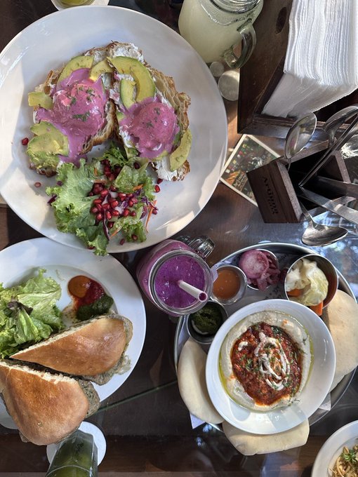 A table is filled with a colorful brunch spread including avocado toast with a pink spread, hummus, salad, bread, and a purple smoothie.