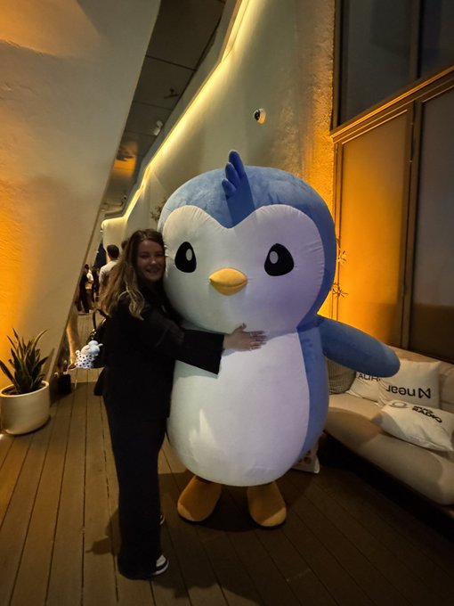 A smiling woman in a black jacket and pants hugs a large inflatable penguin mascot on a wooden deck area at night, with a white sofa visible behind them.