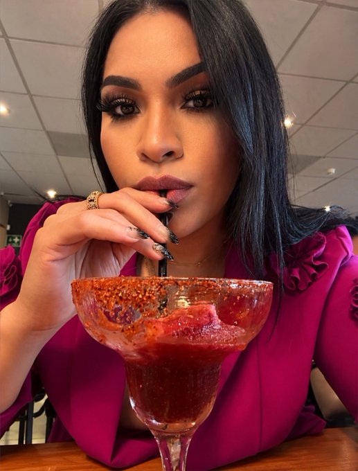 A woman with long dark hair wearing a magenta blouse drinks from a large margarita glass with a chili-salt rim, featuring long decorated nails and a gold ring.