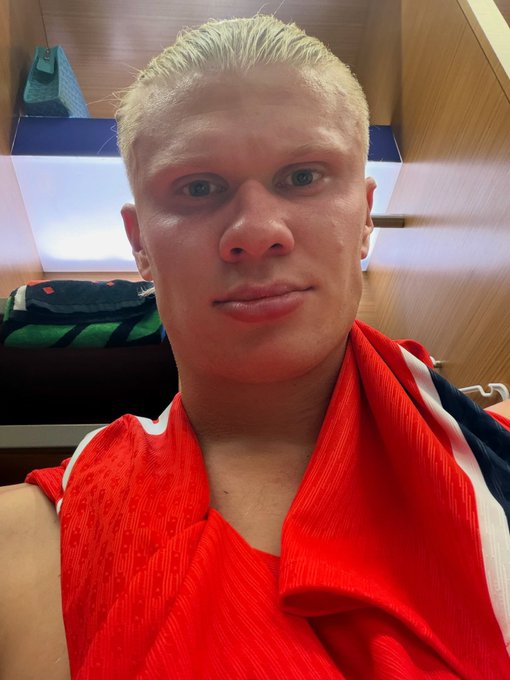 A close-up selfie of a young man with short blonde hair, appearing to be in a locker room, with a red and white sports jersey draped over his shoulder.