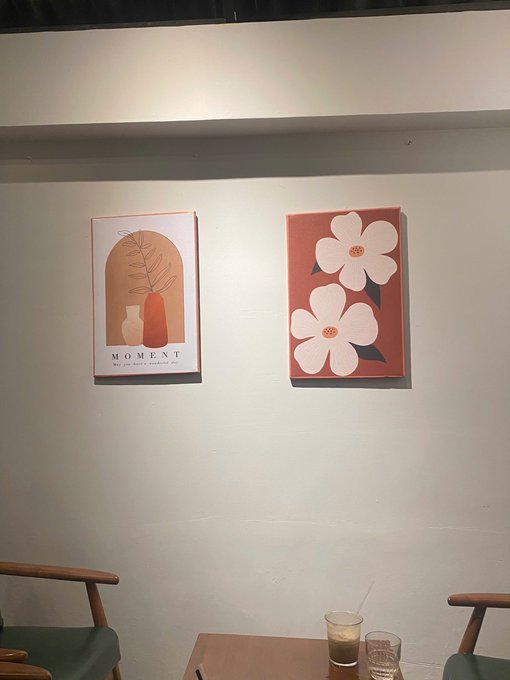 Two framed art prints hang on a white wall in a cafe setting; the left print says 'MOMENT' with vases, while the right features pink flowers on a red background, above wooden chairs and a coffee table.