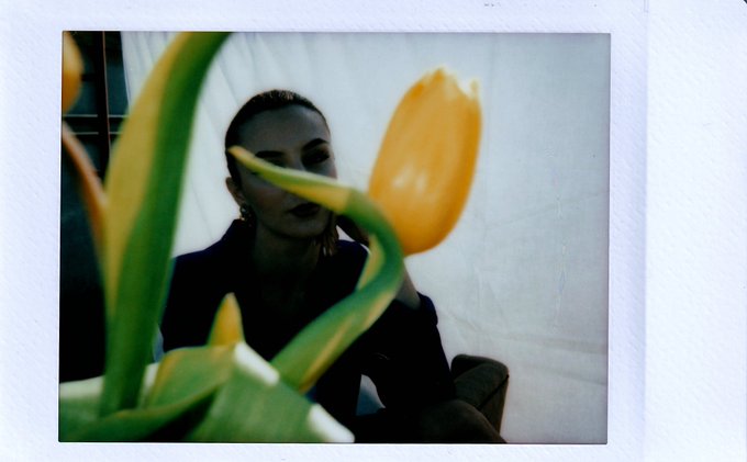 The image features a woman sitting next to a potted plant with yellow flowers, which appears to be a tulip. The background of the photo is white, providing contrast for the colorful elements within the frame. The caption "Gm!Wishing you a weekend in full bloom." suggests that the woman might be celebrating or wishing for a beautiful and vibrant weekend filled with blooming flowers like the one she's sitting next to.