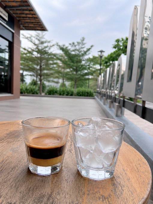 An outdoor scene at a cafe featuring two glasses on a round wooden table in the foreground—one containing a dark espresso drink and the other filled with ice water—with a building and trees in the background.