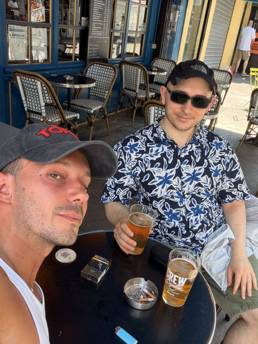 A casual selfie of two men sitting at an outdoor cafe table with beers, cigarettes, and an ashtray; one wears a dark floral shirt and sunglasses, the other a grey baseball cap.