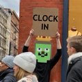 A person holds up a sign reading "CLOCK IN" featuring a green cartoon character, rallying community support for the $CLOCKIN token launch as fans await the rescheduled countdown following weather-related delays.
