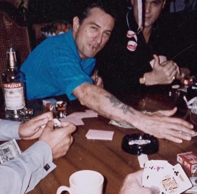 A man in a blue shirt smiles at the camera while playing poker with another player, surrounded by alcohol bottles and playing cards.
