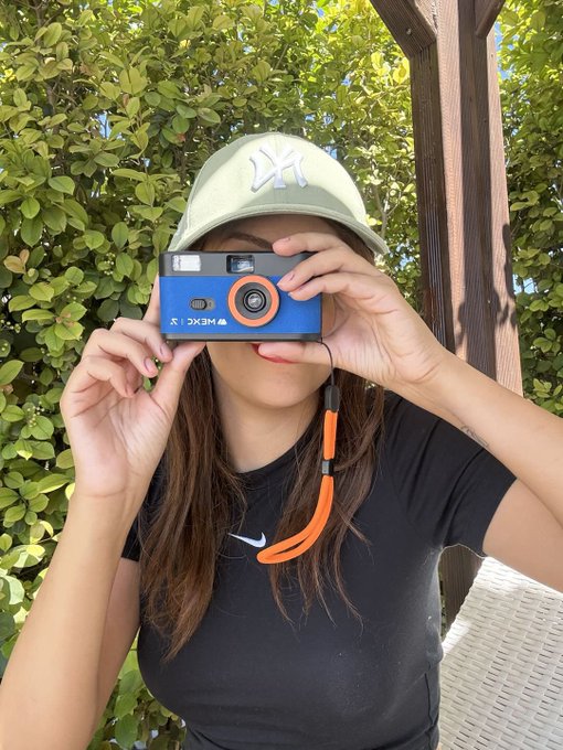 A person wearing a mint green NY Yankees cap holds a blue film camera up to their face while taking a mirror selfie outdoors.