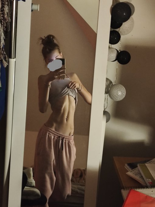 A person takes a mirror selfie wearing a white crop top and pink sweatpants, with their face covered by a gray heart emoji.