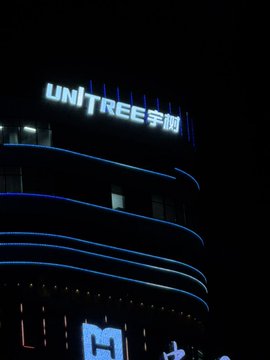 A nighttime photograph of a building illuminated with blue lights featuring curved horizontal lines, topped with a glowing sign that reads 'UNITREE 宇树'.