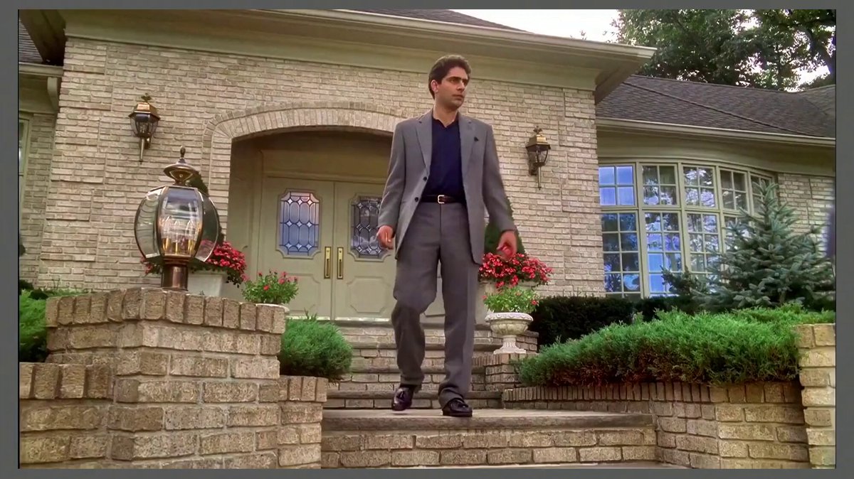 In the scene, there's an actor wearing a suit standing near a brick house with flower boxes on either side of the steps. The man has his hand in his pocket as he poses for the camera. A small television screen is visible behind him, showing something related to a trading game, possibly involving cryptocurrencies or stock markets. The image appears to be from an old TV broadcast and has a vintage look to it.