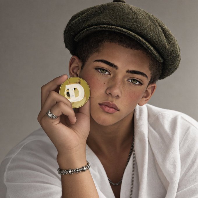 A young person wearing a green newsboy cap and white shirt holds up a yellow circular object marked with a 'D' near their face, accessorized with silver rings and a beaded bracelet.
