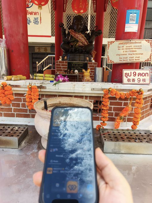 A hand holds a smartphone displaying a cloud wallpaper in front of a small shrine featuring a dark deity statue, marigold garlands, and signs with Thai, Chinese, and English text offering blessings at Wat Mahathat Woramahawihan.