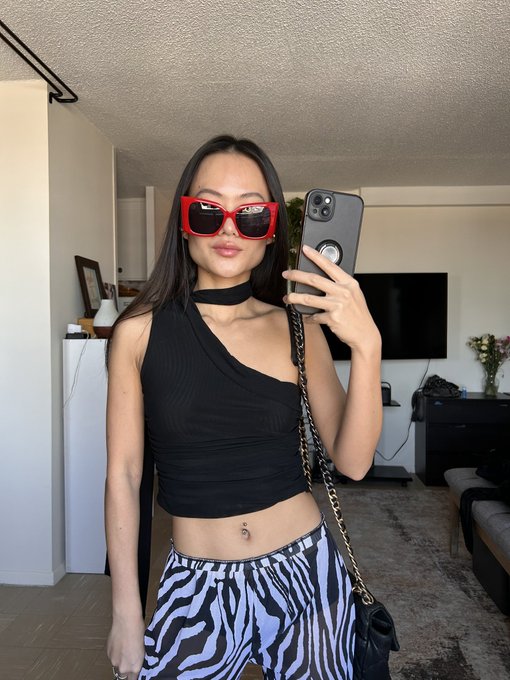 A mirror selfie of a young woman wearing red sunglasses, a black one-shoulder crop top, a choker, and zebra-print pants, with a chain-strap bag in a living room setting.