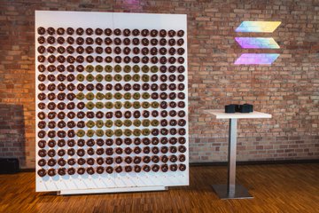 The image features a large white display case filled with rows of chocolate donuts, each one unique and eye-catching. The donuts are arranged in various patterns and designs, creating an impressive visual effect. In addition to the donut wall, there is also a small table placed next to it, possibly for displaying more donuts or serving refreshments during events.

The image includes text that reads "Lambo is cool and all, but did you know we also had other amazing highlights during the @Solana Summit in Germany? - A Solana donut wall - Getting inked with Solana tattoos (real!) - An intense live trading competition - Golfing into the river It was exceptional". This text provides context to the image by mentioning a special event or gathering related to cryptocurrency and donuts.