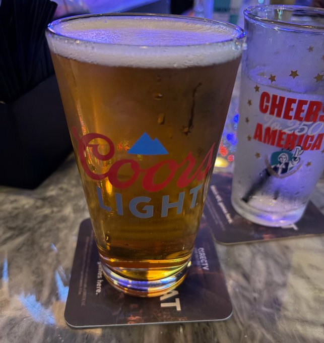 A pint of Coors Light beer sits on a coaster next to a glass reading "Cheers America" in a bar setting, fitting the post's "Beer fund is accelerating" caption celebrating the ticker $GEIGER.