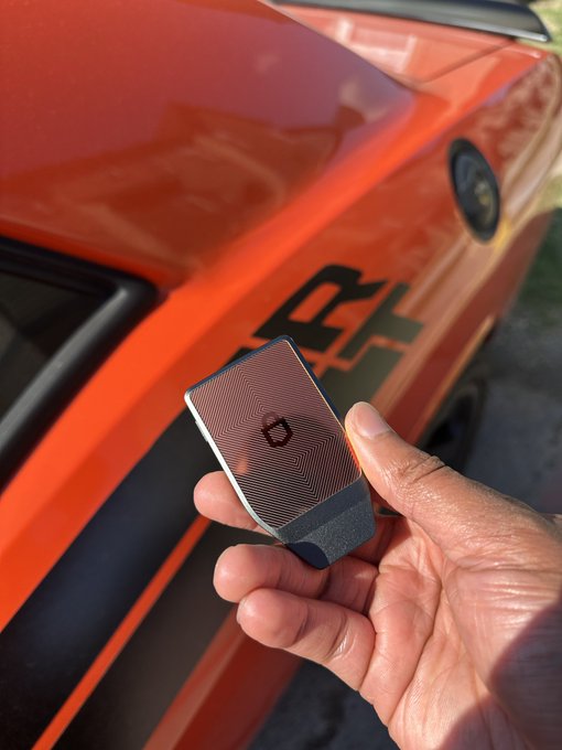 A hand holding a small dark gray hardware crypto wallet with a logo in front of an orange sports car (appears to be a Chevrolet Corvette with 'C4' or similar badge visible).