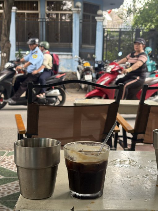 A glass of iced black coffee and a metal cup sit on a table in the foreground, overlooking a busy street scene with motorbikes and pedestrians in the background.