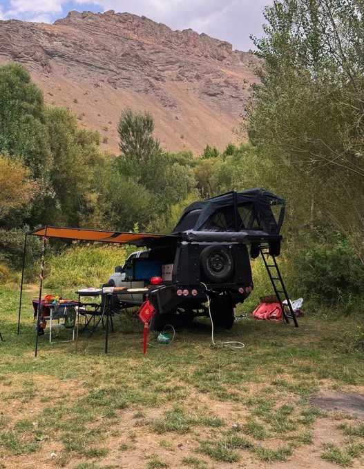 An off-road camping setup featuring a black 4x4 truck equipped with a rooftop tent, awning, and ladder, parked on grass with a rocky mountain backdrop.