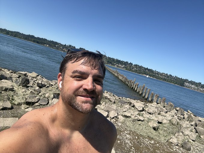 A shirtless man with an earbud takes a selfie on a rocky shoreline with calm blue water, a wooden breakwater, and forested hills in the background.