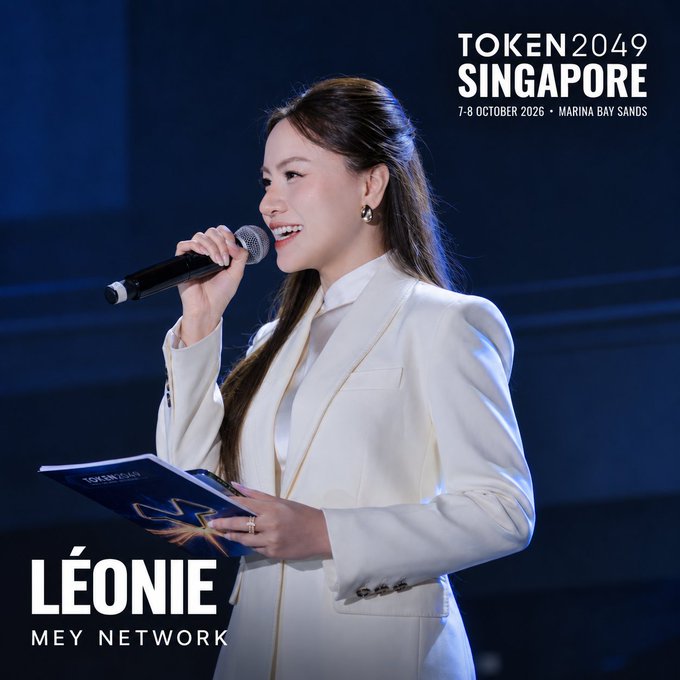 A woman in a white blazer holds a microphone and speaks at TOKEN2049 Singapore event (7-8 October 2026, Marina Bay Sands), identified as Léonie from MEY Network.
