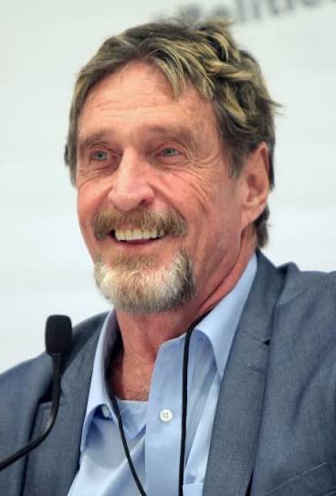 A photograph of an older man with a gray beard and mustache wearing a gray blazer over a light blue shirt, smiling while speaking at a microphone.