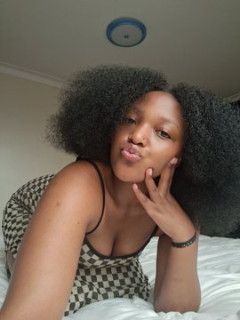 A young woman with curly dark hair taking a selfie indoors, wearing a black and white checkered top, with her hand resting near her face.