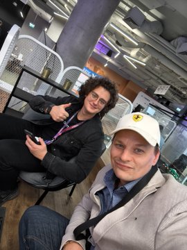 Two men taking a selfie indoors, one wearing glasses and a dark jacket, the other in a light jacket and beige cap with a yellow logo.