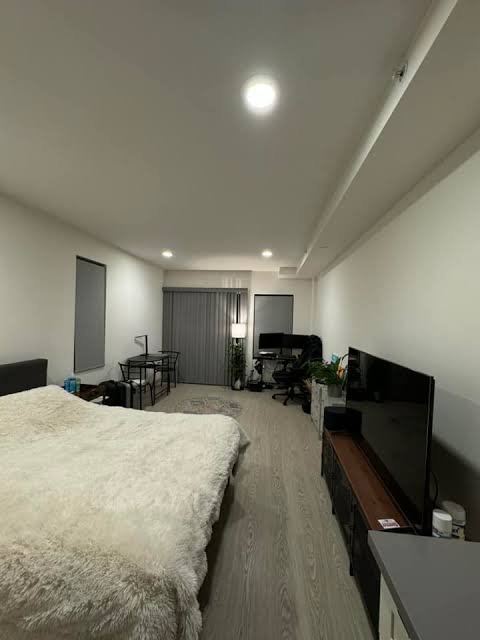 A modern bedroom interior featuring a bed with white fluffy bedding, a wall-mounted TV on a wooden console, a desk setup with a computer monitor, and recessed ceiling lighting.