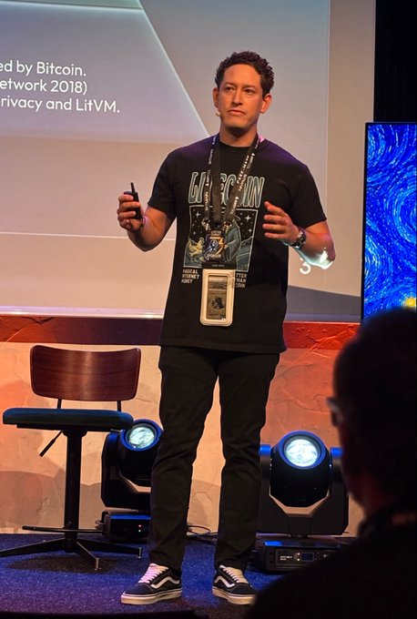 A presenter stands on stage at the Litecoin Summit, wearing a "84 Million Magical Internet Money" t-shirt and an LTCG 2026 Margaret "God Mode" VIP lanyard, while holding a clicker and addressing the audience beside a slide mentioning Bitcoin, Network 2018, and LitVM.