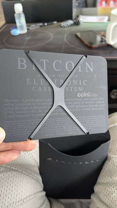 The image shows a person holding up a black wallet with a Bitcoin symbol on it. The wallet has an x-shaped design that allows for easy opening and closing of the wallet. The person's hand is positioned over the wallet, showcasing its features. The background includes a desk with various items such as a cell phone, a cup, and a computer mouse.