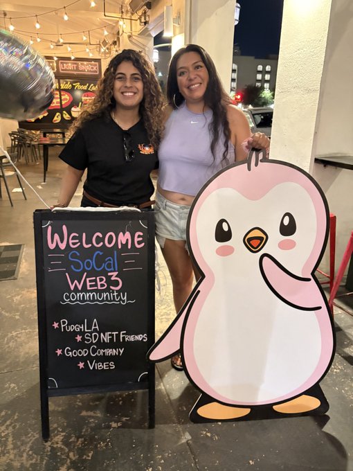 Two smiling women standing next to a welcome sign for the 'SoCal WEB3 community' featuring 'Pudgy LA', 'SD NFT Friends', 'Good Company', and 'Vibes', alongside a penguin cutout.