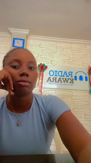 A young woman in a light blue t-shirt poses for a selfie in what appears to be a radio station studio, with a mirrored "RADIO KWARA" sign visible in the background, aligning with her caption about serving at the station. She wears a delicate necklace and rests her chin on her hand, giving a calm, thoughtful expression.