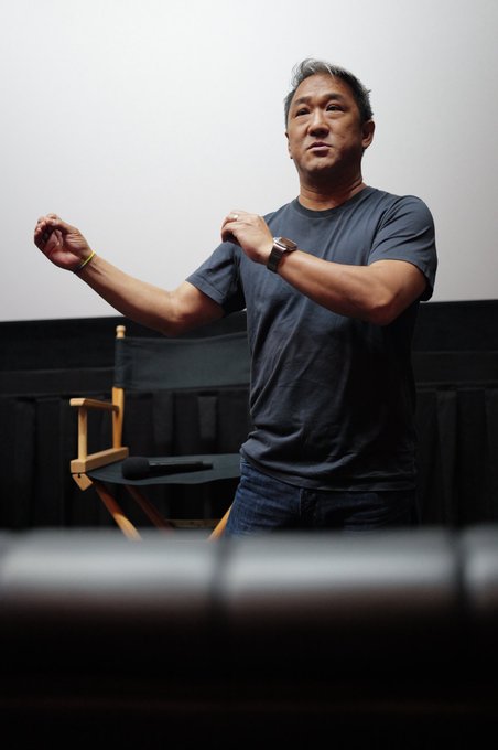 A man in a dark t-shirt stands in a director's chair pose, gesturing with his hands in what appears to be a studio setting.