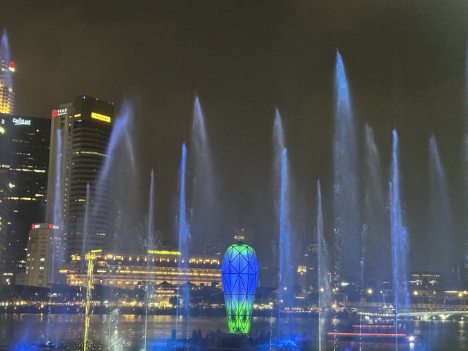 A nighttime cityscape featuring an illuminated water fountain show with blue-lit water jets, the colorful lotus-shaped ArtScience Museum, and surrounding skyscrapers including buildings labeled 'CapitaLand' and 'HSBC'.