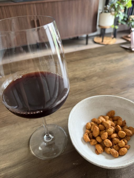 A glass of red wine sits on a wooden table next to a white bowl filled with roasted almonds, with wooden cabinets and a plant visible in the background.