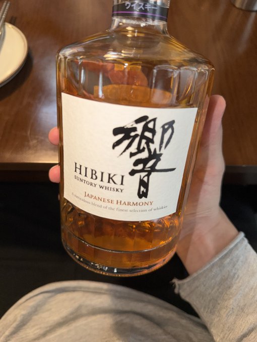A person holds a nearly empty bottle of Hibiki Japanese Harmony Suntory Whisky at a dinner table, fitting the Bitcoin maximalist theme since "HODL"—a crypto mantra about never selling—originated from a misspelling of "hold" in a 2013 Bitcoin forum post about whiskey during a market crash.