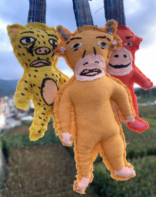 Three handmade felt animal plushies—a yellow leopard, an orange horse-like creature, and a pink figure—hang against a blurred green and blue background.
